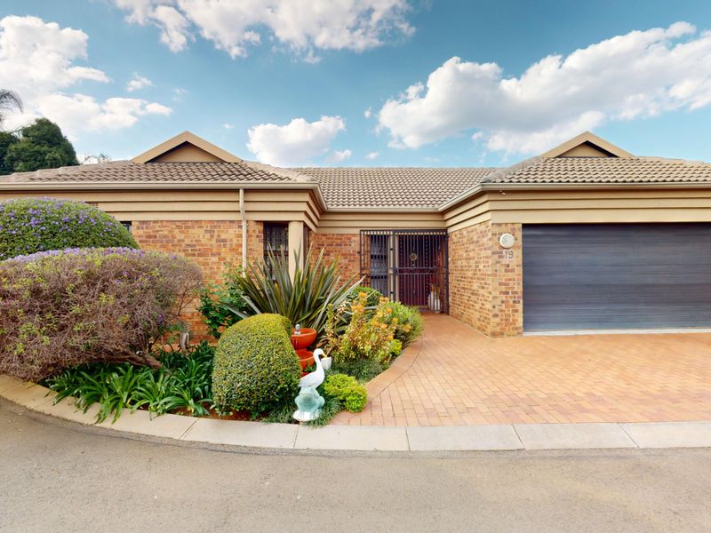 Front elevation of a well-maintained brick house featuring a paved driveway, double garage, and established garden with security gate entrance.