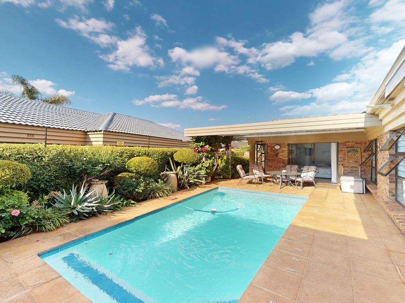 A well-maintained outdoor pool area featuring a tiled patio, covered entertainment space, and established garden hedging.