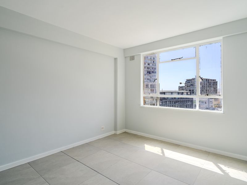 A bright, empty bedroom featuring neutral tiled flooring and a large window with urban views.