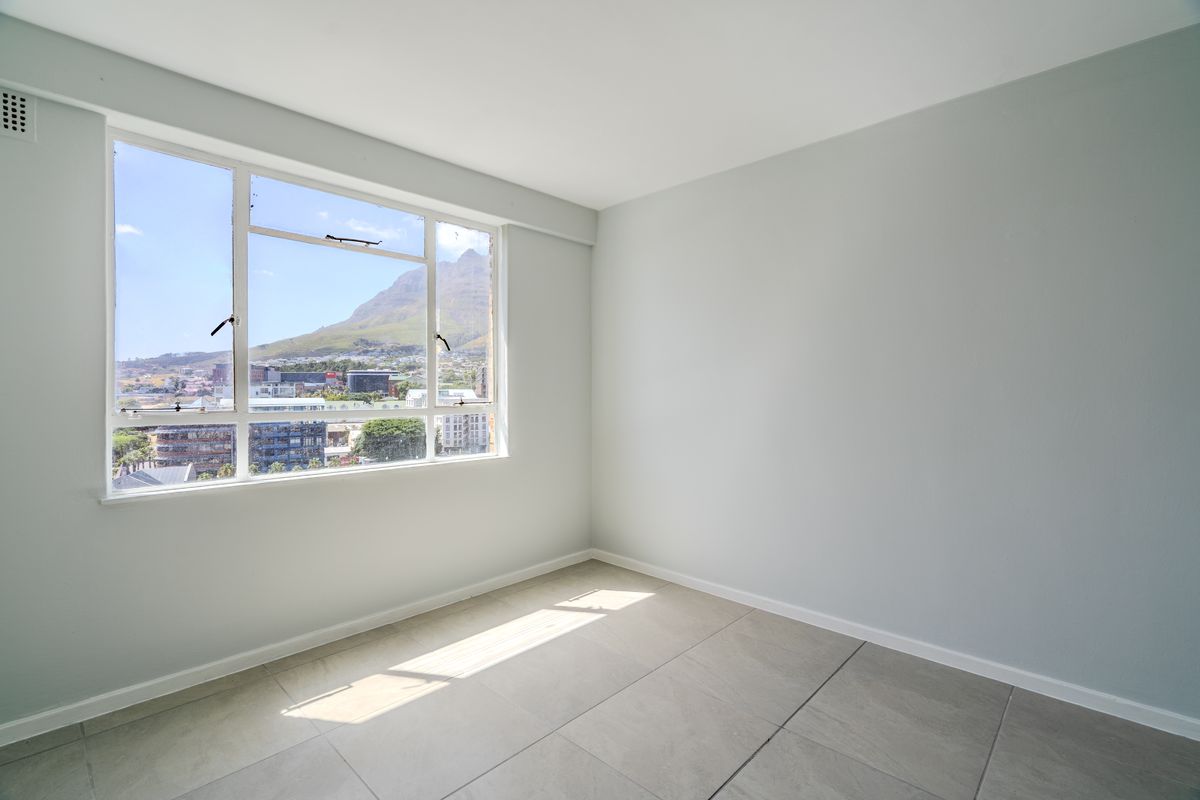 A bright, empty bedroom featuring large windows with mountain and city views, neutral wall paint, and clean tiled flooring.