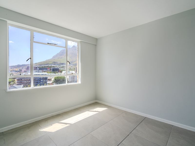 A bright, empty bedroom featuring large windows with mountain and city views, neutral wall paint, and clean tiled flooring.