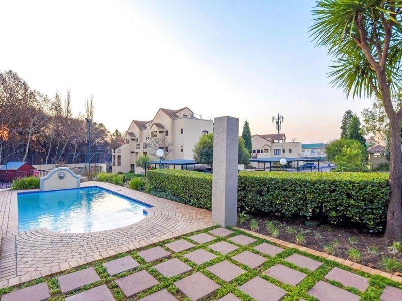 A well-maintained communal swimming pool area featuring a paved deck, decorative water feature, and established garden hedging within an apartment complex.