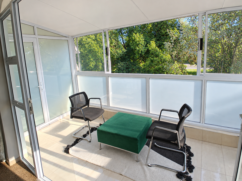 A bright, enclosed balcony featuring tiled flooring and large aluminium-framed windows, offering a pleasant view of the surrounding greenery.