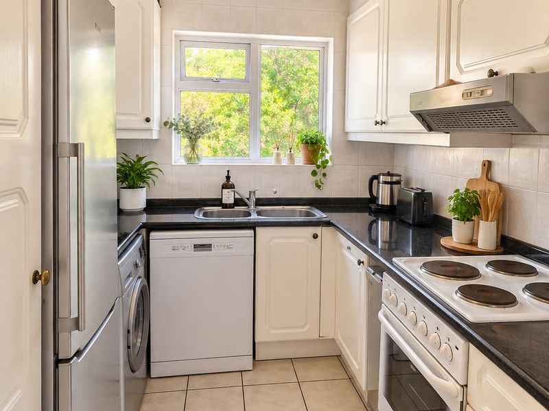 A well-maintained galley kitchen featuring white cabinetry, dark countertops, integrated appliances, tiled flooring, and a window providing natural light.