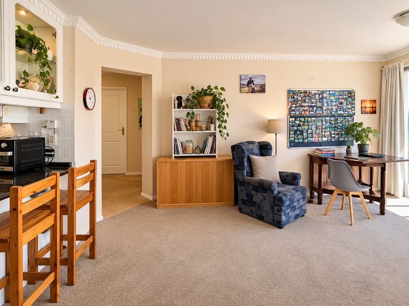 An open-plan living and kitchen area featuring neutral carpeting, crown molding, and a functional kitchen corner with granite countertops and built-in cabinetry.