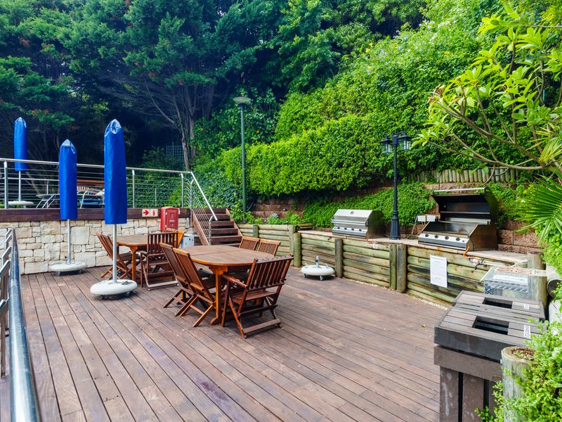Outdoor entertainment deck featuring timber flooring, stainless steel balustrades, and a built-in gas braai area set against a landscaped garden backdrop.