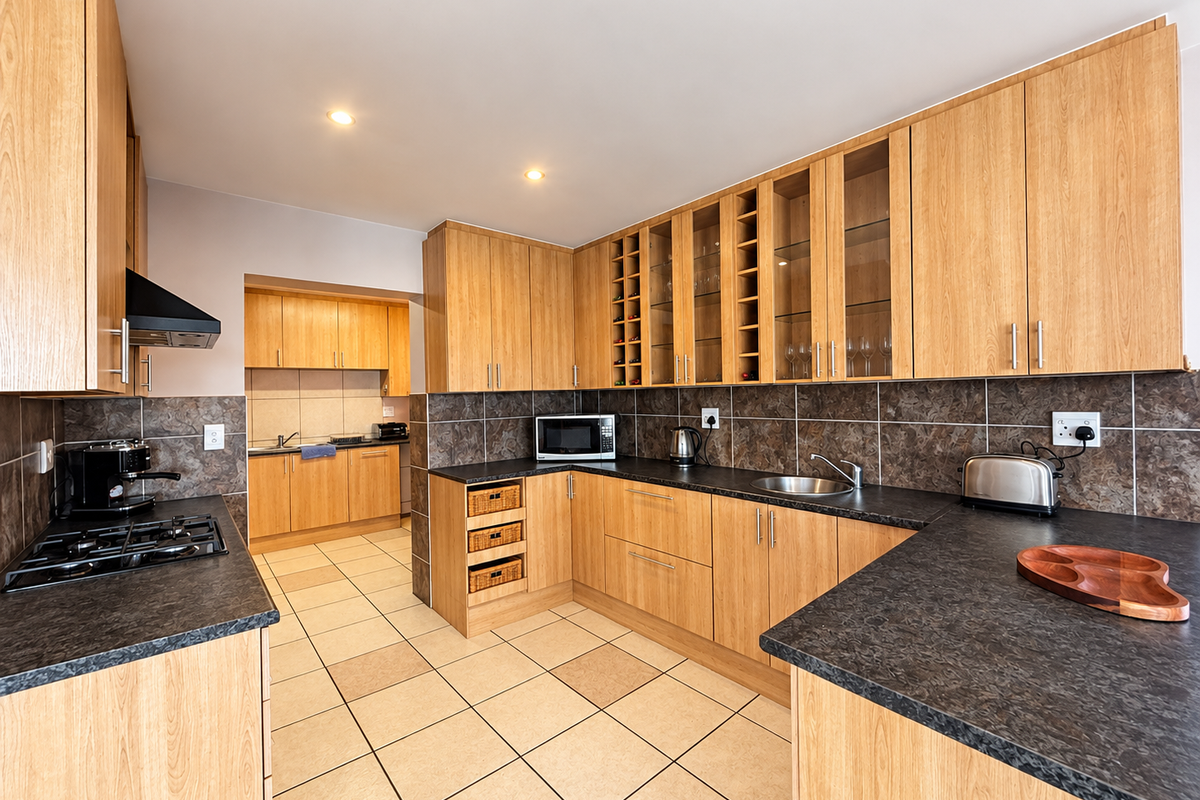 A functional kitchen featuring light wood cabinetry, dark laminate countertops, a gas hob with extractor, and tiled flooring with an adjoining scullery area.