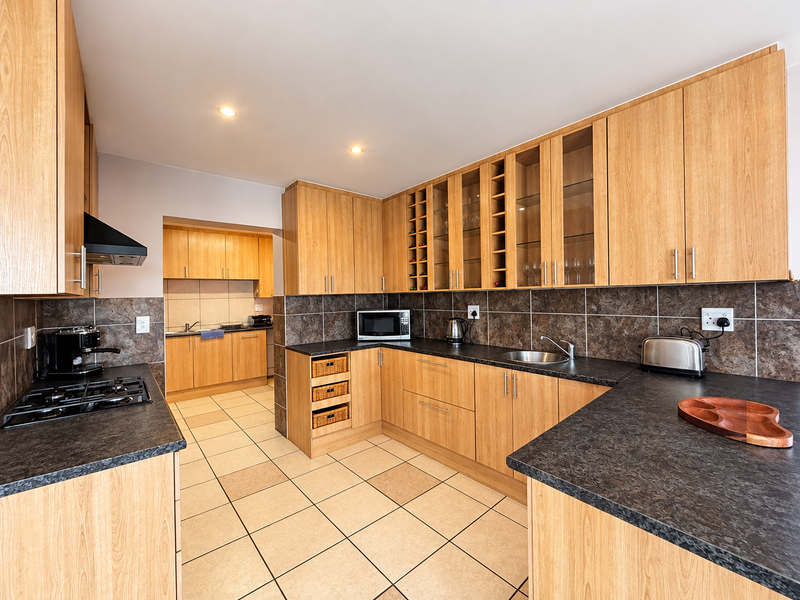 A functional kitchen featuring light wood cabinetry, dark laminate countertops, a gas hob with extractor, and tiled flooring with an adjoining scullery area.