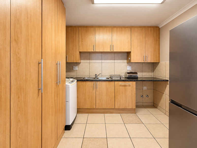 Functional scullery featuring light wood-look cabinetry, dark laminate countertops, tiled backsplash, and dedicated plumbing connections for laundry appliances.