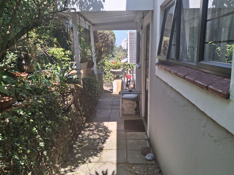 A narrow, covered paved walkway along the side of the cottage, featuring aluminium window frames and a brick sill, leading to the rear garden area.