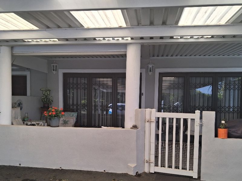 A covered patio area featuring a corrugated roof with translucent panels, concrete columns, and secure sliding glass doors with metal security gates.