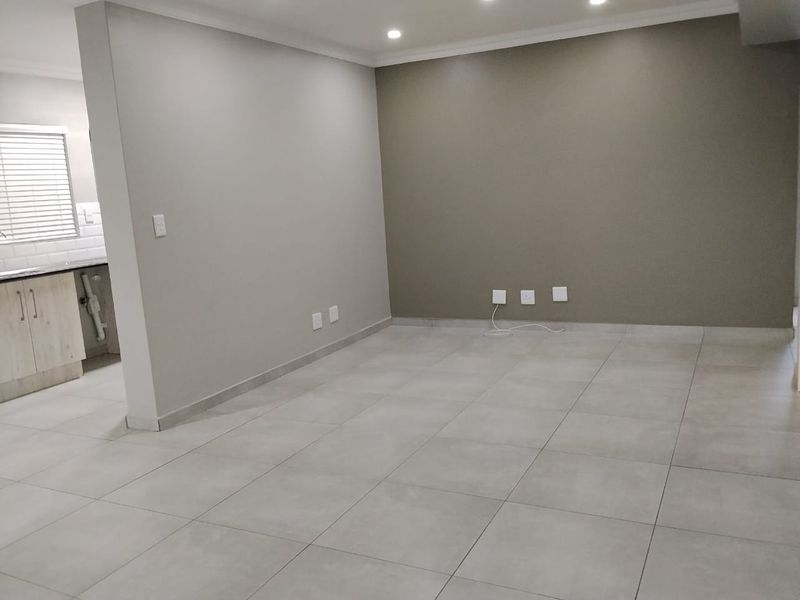 An open-plan living area featuring neutral tiled flooring, recessed lighting, and a partial view into a modern kitchen with built-in cabinetry.