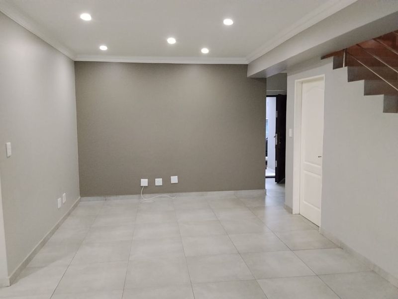 An empty, well-maintained living area featuring neutral tiled flooring, recessed downlighting, and a staircase leading to the upper level.