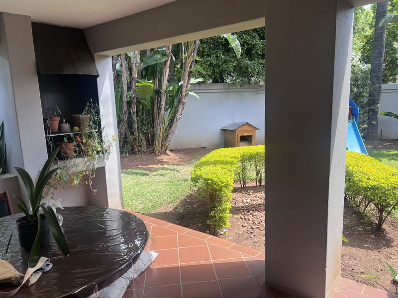 A covered patio area featuring a built-in braai and tiled flooring, overlooking a landscaped garden with mature trees and lawn.