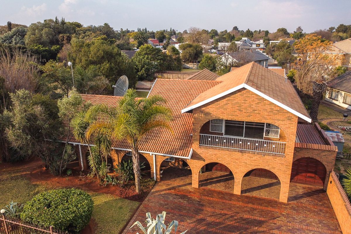 Aerial view of a double-story brick house featuring a tiled roof, arched ground-level portico, upper balcony, and a paved driveway set within a landscaped garden.
