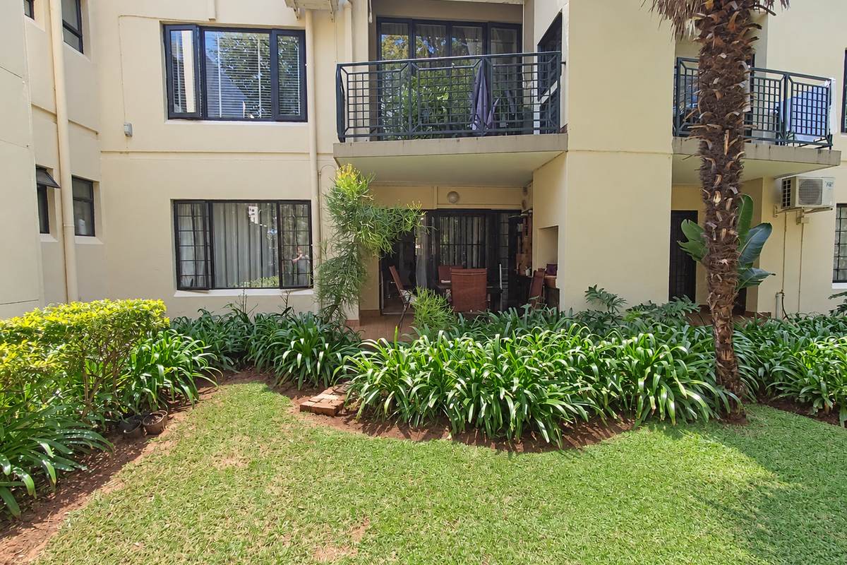 Rear view of the apartment building featuring a ground floor patio, upper level balcony with metal railings, and a well-maintained private garden area.