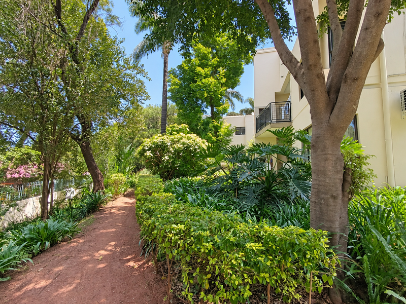 A lush, well-maintained garden area featuring mature trees, manicured hedges, and a pathway alongside the apartment building exterior.