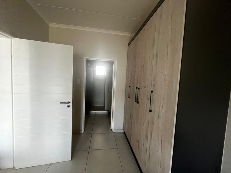 A clean, empty bedroom featuring modern built-in cupboards with wood-grain finish and neutral tiled flooring, leading to an adjoining bathroom.