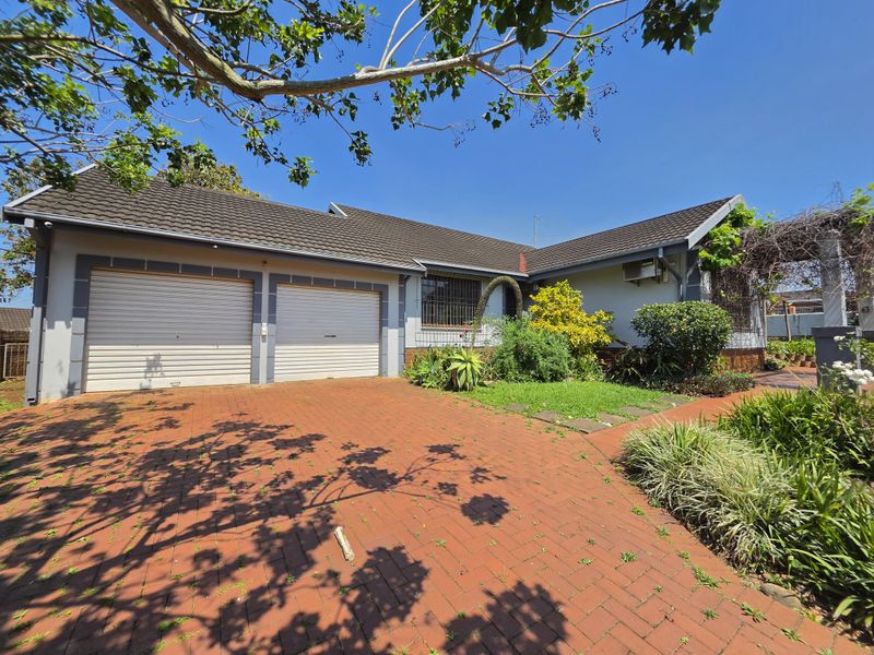 Front exterior view of a single-story house featuring a double garage with roll-up doors, a brick-paved driveway, and established garden landscaping.