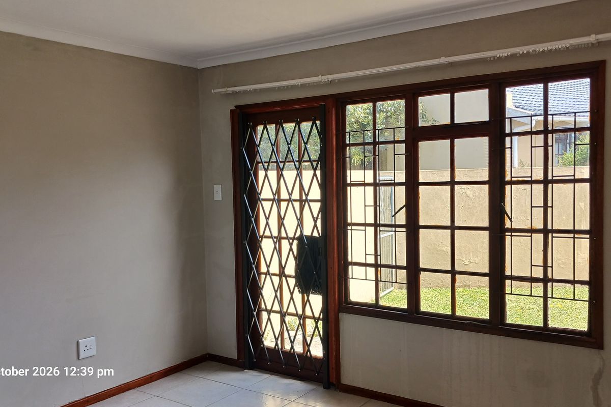 An empty living room featuring tiled flooring, wooden window frames, and a security gate leading to the exterior garden area.