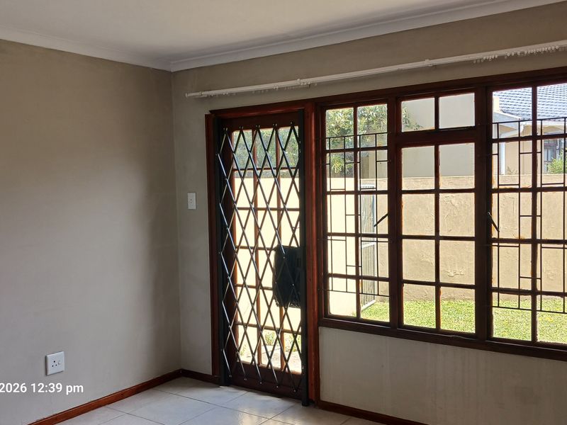 An empty living room featuring tiled flooring, wooden window frames, and a security gate leading to the exterior garden area.