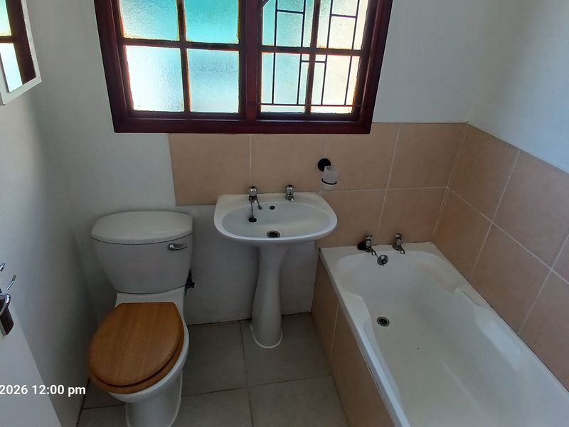 A functional bathroom featuring a pedestal basin, toilet, and built-in bathtub with tiled splashbacks and a window with security bars.