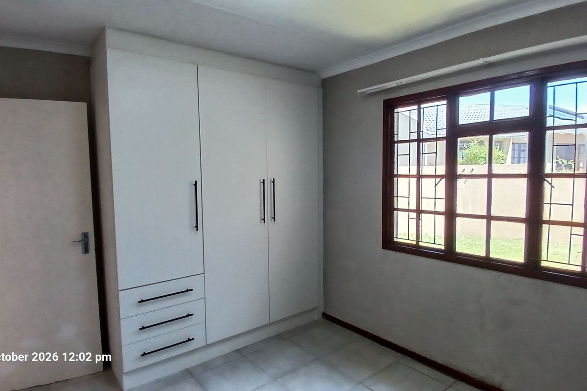 A bright, empty bedroom featuring built-in white cupboards, tiled flooring, and a window fitted with security bars.