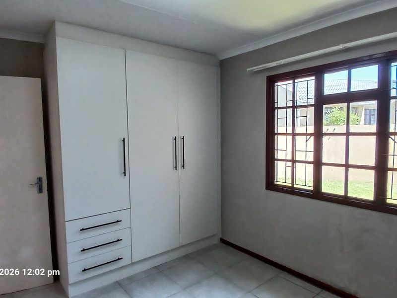 A bright, empty bedroom featuring built-in white cupboards, tiled flooring, and a window fitted with security bars.