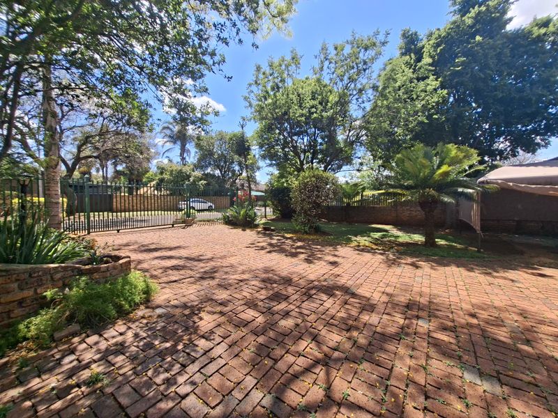 A spacious front exterior area featuring a brick-paved driveway, secure metal gate, and established garden with mature trees and boundary walling.