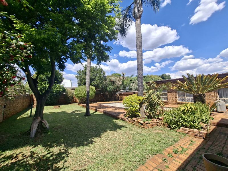 A spacious backyard featuring a swimming pool, brick paved patio area, and established garden landscaping with mature trees and boundary walls.