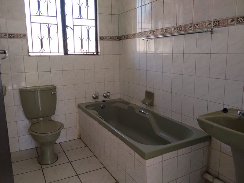 A dated bathroom featuring a full-sized bathtub, pedestal basin, and toilet, finished with wall-to-floor tiling and security bars on the window.