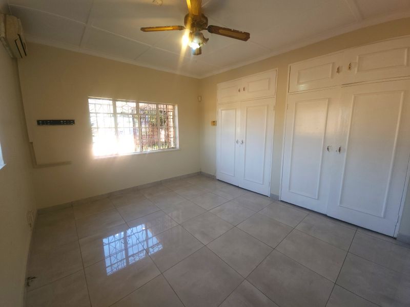 A spacious, empty bedroom featuring neutral tiled flooring, built-in cupboards, a ceiling fan, and an air conditioning unit.