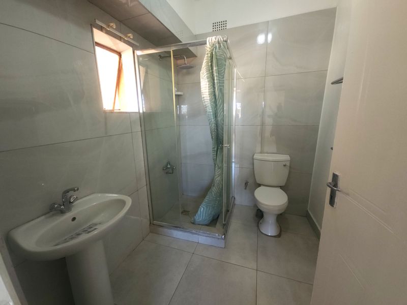 A clean, modern bathroom featuring a pedestal basin, a glass-enclosed shower, and a toilet, finished with neutral wall and floor tiles.