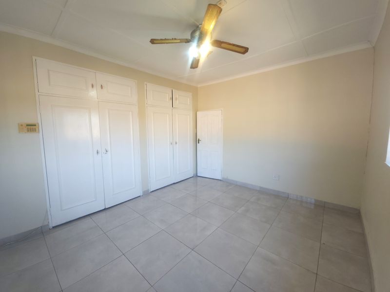 A spacious, empty bedroom featuring neutral tiled flooring, built-in cupboards, and a ceiling fan, offering a clean canvas for personalization.