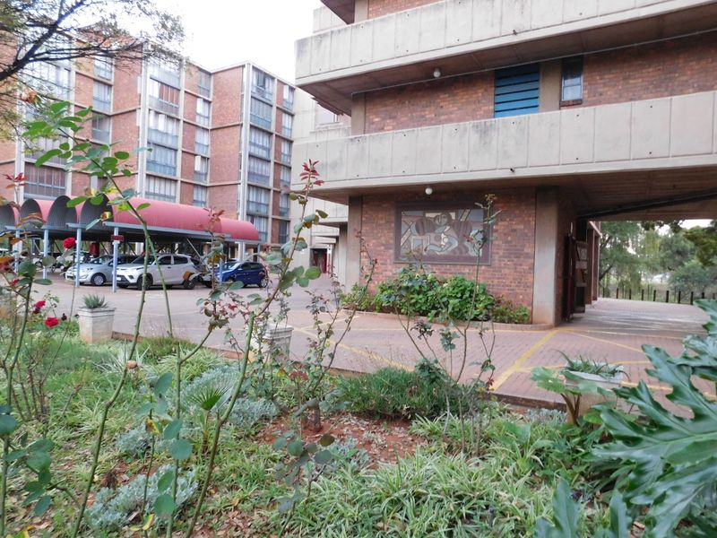 Exterior view of a brick apartment complex featuring covered parking, concrete balconies, and a paved driveway area with established garden beds.