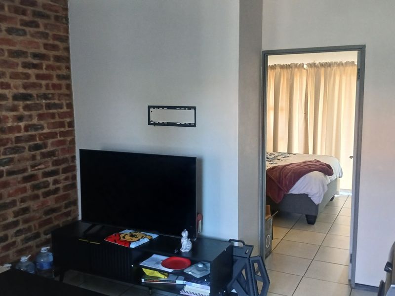 Living area featuring an exposed brick feature wall and tiled flooring, with an adjacent bedroom visible through the doorway.