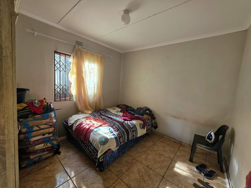 A bedroom featuring tiled flooring, a window with security gate, and basic ceiling lighting. The room requires cosmetic updates to the wall finishes.