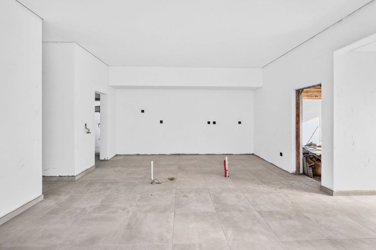An unfinished, open-plan living area featuring large format floor tiles, exposed plumbing, and bare walls, currently in a state of renovation.