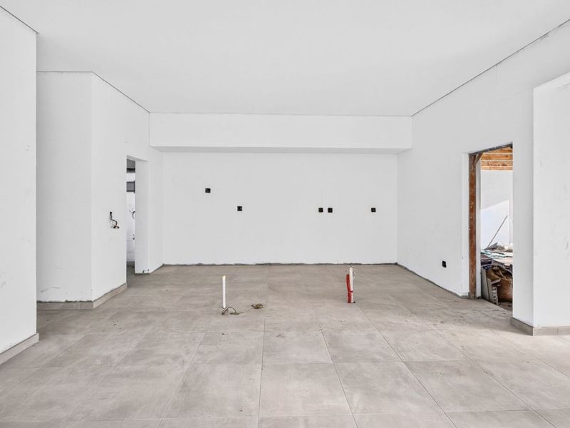 An unfinished, open-plan living area featuring large format floor tiles, exposed plumbing, and bare walls, currently in a state of renovation.