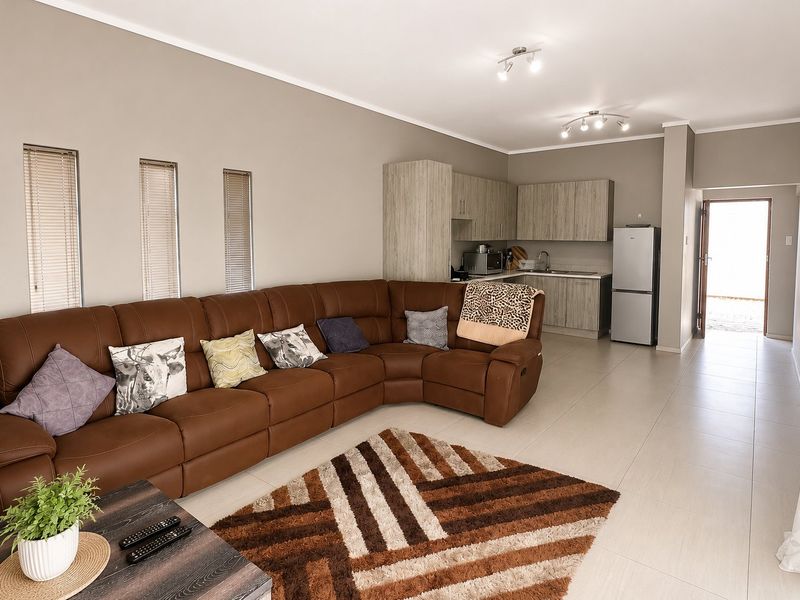 Open-plan living area featuring tiled floors, neutral walls, and a functional kitchen with built-in cabinetry and sink, leading to an exterior entrance.