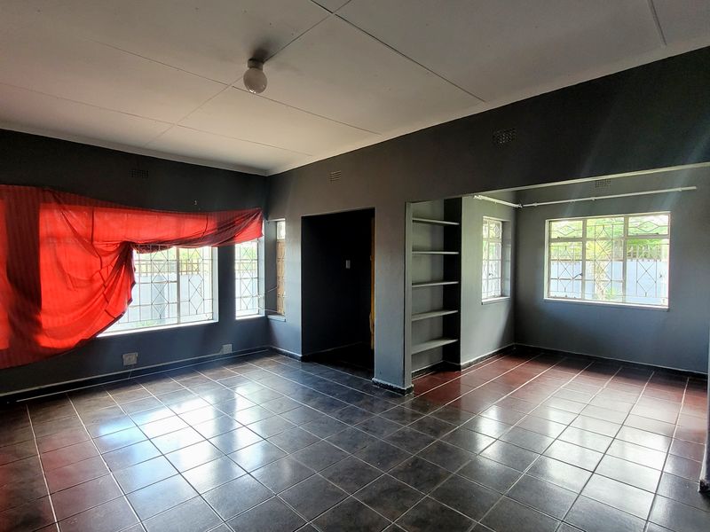 An empty open-plan living area featuring dark tiled floors, built-in shelving, and windows fitted with security bars, awaiting a fresh update.