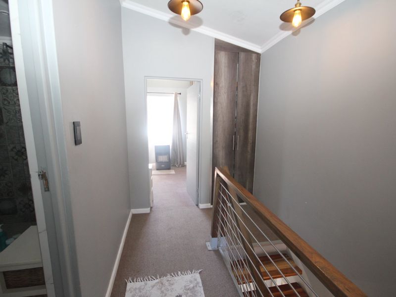A well-maintained hallway featuring a staircase with a timber handrail, stainless steel balustrade, built-in storage cupboard, and modern pendant lighting.