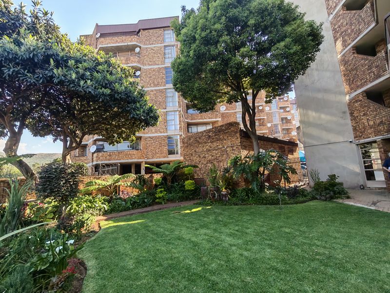 A well-maintained rear garden area featuring mature trees and lush landscaping, set against the face brick facade of a multi-story residential apartment building.
