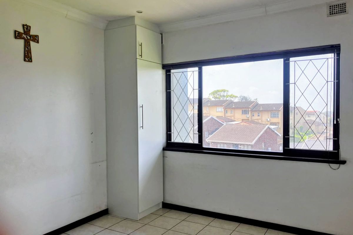 A bright, empty bedroom featuring tiled flooring, a built-in cupboard, and large windows with security bars, offering a view of the surrounding townhouse complex.