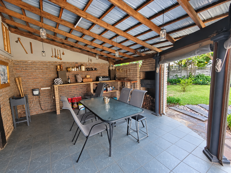 Covered outdoor patio featuring a built-in brick braai, exposed timber roof beams, and tiled flooring, overlooking a private garden.