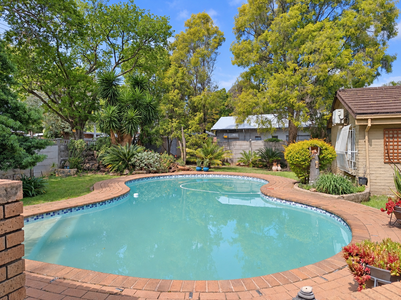 A private backyard featuring a kidney-shaped swimming pool surrounded by brick paving, established trees, and landscaped garden beds.