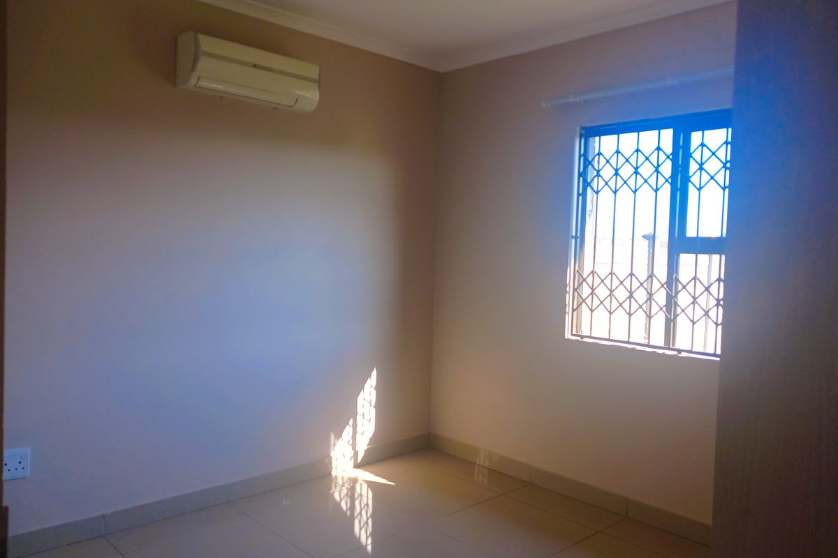 A bright, empty bedroom featuring tiled flooring, a wall-mounted air conditioning unit, and a window fitted with a security gate.