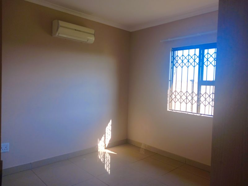 A bright, empty bedroom featuring tiled flooring, a wall-mounted air conditioning unit, and a window fitted with a security gate.