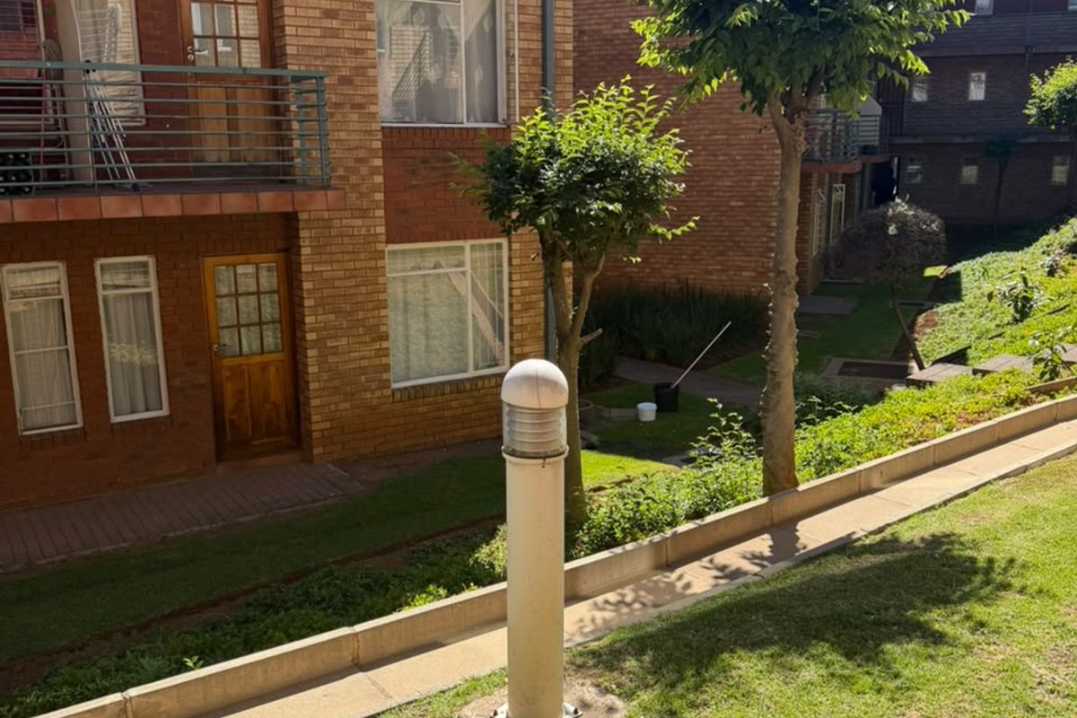 A view of the apartment complex rear exterior featuring brick facade, private balconies with metal railings, and a landscaped garden area with paved pathways.