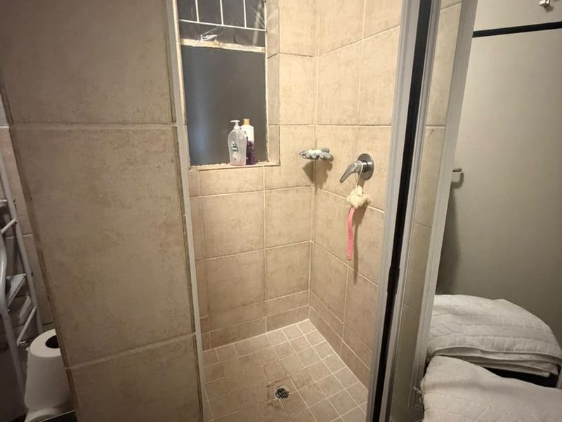 A compact bathroom shower area featuring beige wall and floor tiles, a built-in shower mixer, and a window with security bars.